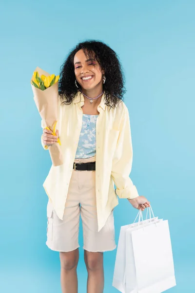 Pleased african american woman in stylish clothes posing with yellow tulips and shopping bags isolated on blue — Stock Photo