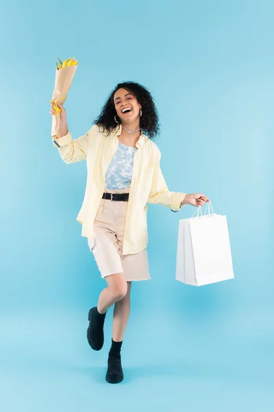 Full length of overjoyed african american woman in shorts and black boots posing with yellow tulips and shopping bags on blue — Stock Photo