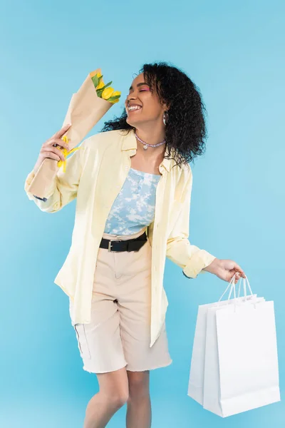 Mujer afroamericana de moda con tulipanes amarillos y bolsas de compras sonriendo con los ojos cerrados aislados en azul - foto de stock