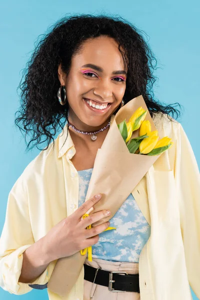Feliz y elegante mujer afroamericana sosteniendo tulipanes amarillos aislados en azul - foto de stock