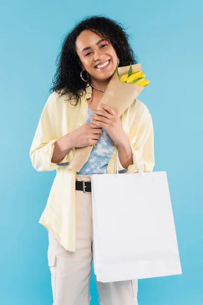 Mujer afroamericana feliz con bolsas de compras y tulipanes amarillos mirando a la cámara aislada en azul - foto de stock