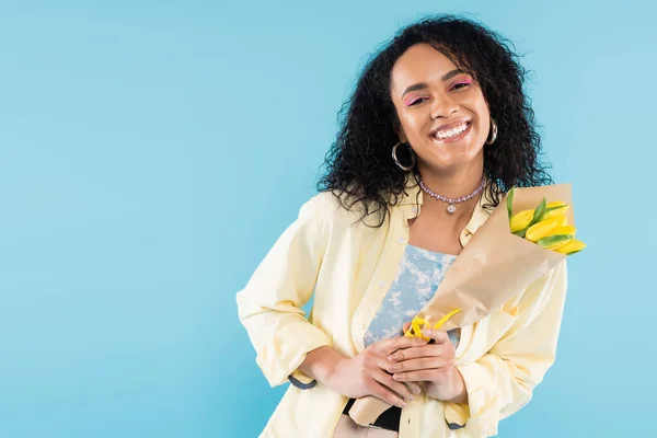 Trendy african american woman in hoop earrings and beads holding yellow tulips and smiling at camera isolated on blue — Stock Photo