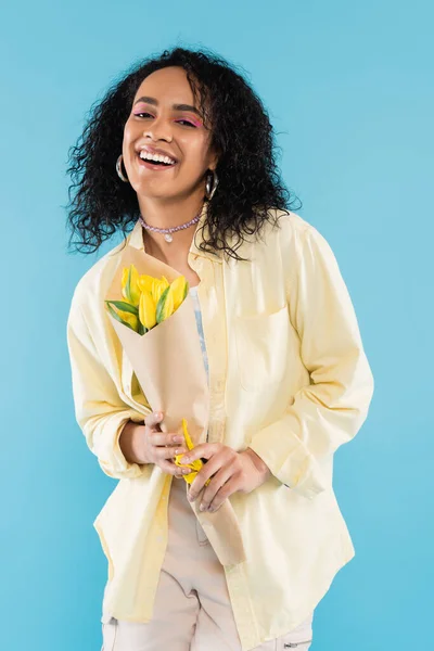 Mujer afroamericana feliz en camisa elegante que sostiene los tulipanes amarillos aislados en azul - foto de stock