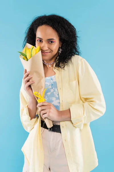 Heureux afro-américain femme en vêtements à la mode tenant bouquet de tulipes jaunes isolé sur bleu — Photo de stock
