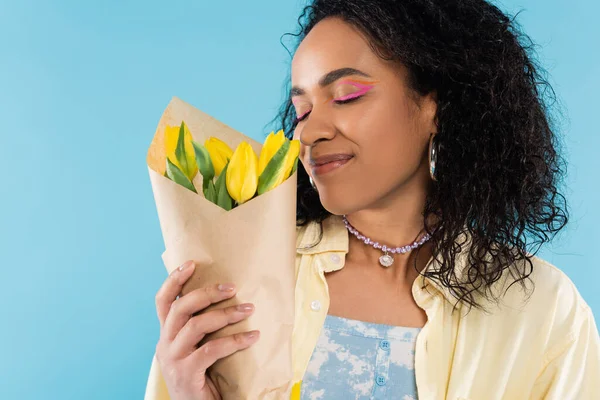 Piacere donna afro-americana con gli occhi chiusi godendo sapore di tulipani gialli isolati su blu — Foto stock
