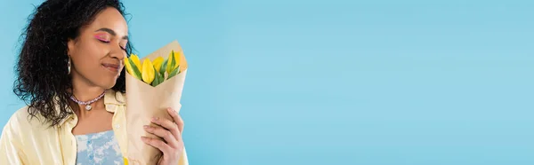 Pleased african american woman enjoying flavor of yellow tulips isolated on blue, banner — Stock Photo