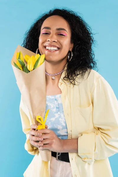 Donna afroamericana trendy e felice con capelli ricci bruna che tengono tulipani gialli e guardando la fotocamera isolata sul blu — Foto stock