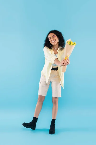 Longitud completa de la alegre mujer afroamericana en pantalones cortos posando con tulipanes amarillos sobre fondo azul - foto de stock