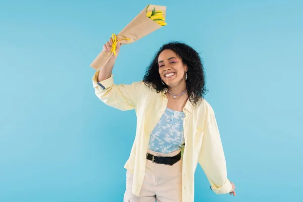 Радісна і модна афроамериканка посміхається на камеру, тримаючи жовті тюльпани ізольовані на синьому — стокове фото