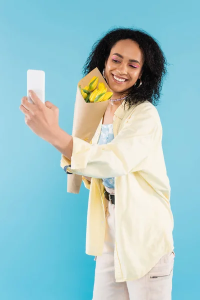 Cheerful african american woman with yellow tulips taking selfie on mobile phone isolated on blue — Stock Photo