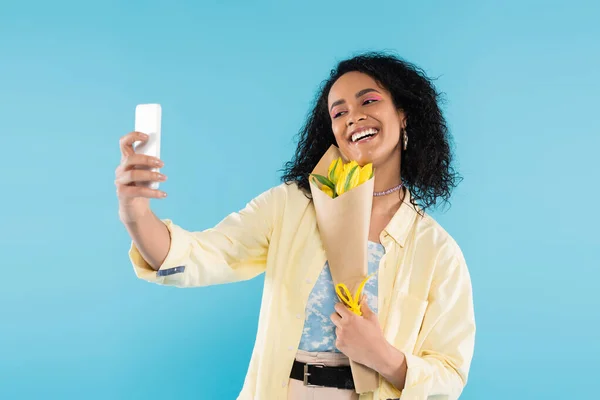 Mulher americana africana alegre e elegante com tulipas amarelas tomando selfie no smartphone isolado no azul — Fotografia de Stock