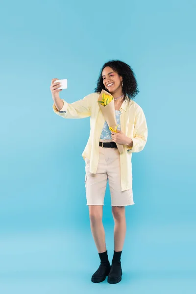 Comprimento total de mulher americana africana sorridente com tulipas amarelas tirando selfie no telefone celular em azul — Fotografia de Stock