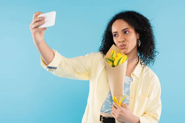 Brunette african american woman with yellow tulips pouting lips and taking selfie on cellphone isolated on blue — Stock Photo