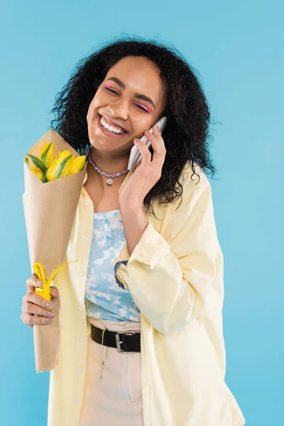 Задоволена афроамериканська жінка з жовтими тюльпанами і закритими очима розмовляє на мобільному телефоні ізольовано на синьому — стокове фото