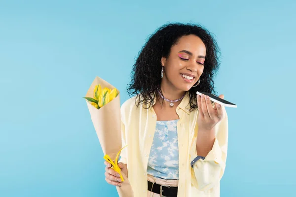 Mujer afroamericana positiva con tulipanes amarillos que envían mensaje de voz en un teléfono inteligente aislado en azul - foto de stock