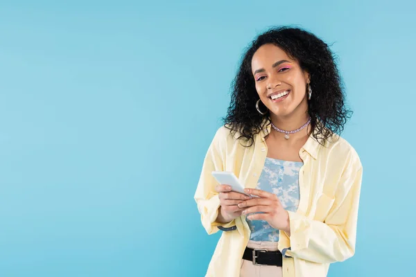 Радісна афро-американська жінка в стильному одязі зі зі смартфоном і дивиться на фотоапарат, ізольований на синьому — стокове фото