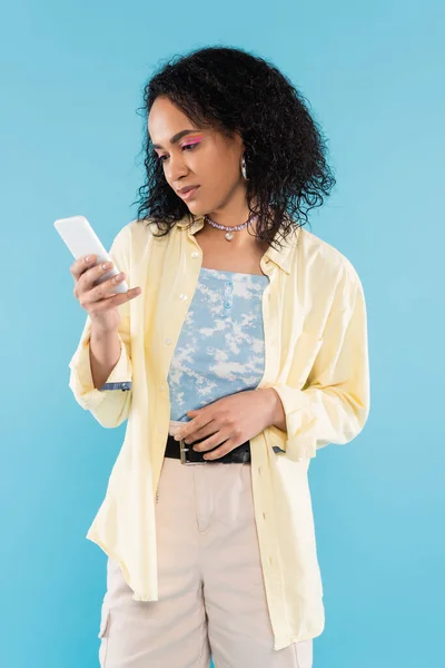 Jovem e moderna mulher americana africana com cabelo encaracolado olhando para smartphone isolado em azul — Fotografia de Stock
