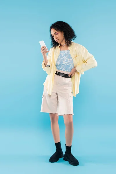 Pleine longueur de femme afro-américaine élégante en short et bottes noires regardant smartphone sur fond bleu — Photo de stock