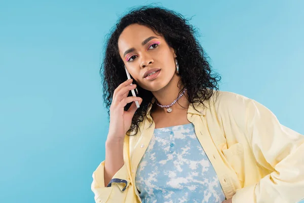 Morena afroamericana americana mujer en camisa amarilla hablando por teléfono móvil y mirando hacia otro lado aislado en azul - foto de stock