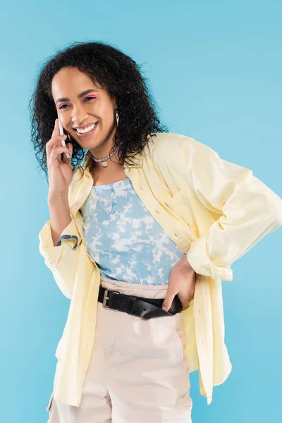 Donna afroamericana spensierata ed elegante che parla sullo smartphone mentre è in piedi con mano sull'anca isolata sul blu — Foto stock