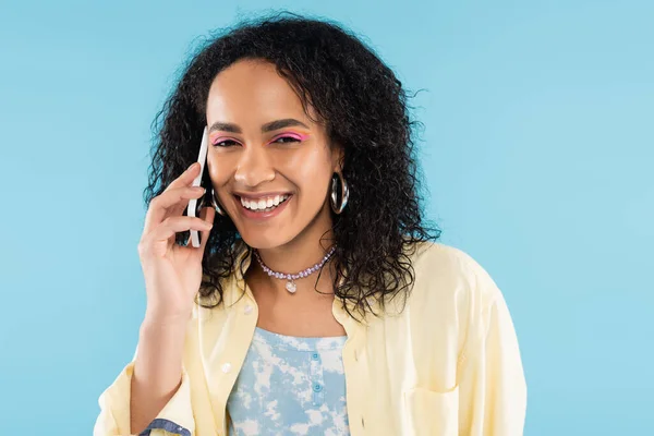 Overjoyed african american woman with wavy brunette hair talking on mobile phone isolated on blue — Stock Photo