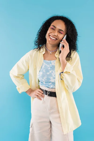 Femme afro-américaine à la mode et heureuse debout avec la main sur la hanche et parler sur un téléphone portable isolé sur bleu — Photo de stock