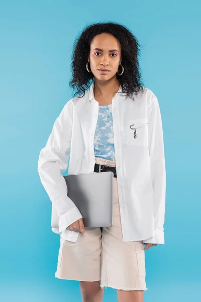 Brünette afrikanisch-amerikanische Frau in weißem Hemd mit Laptop und Blick in die Kamera isoliert auf blau — Stockfoto