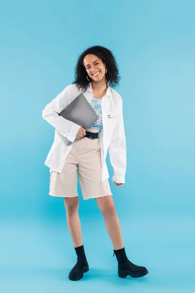 Unbekümmerte Afroamerikanerin in weißen Shorts und schwarzen Stiefeln posiert mit Laptop auf blauem Hintergrund — Stockfoto