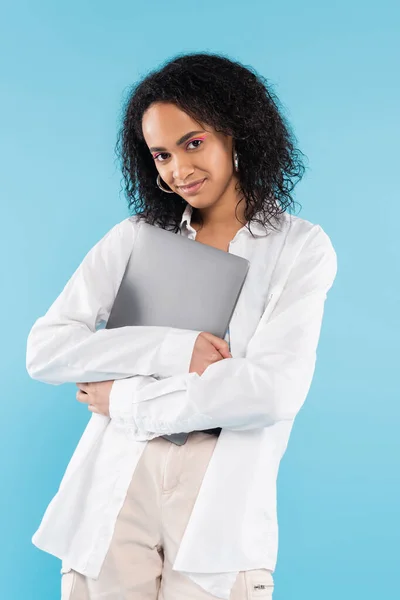 Щаслива афроамериканська жінка в білій сорочці, що стоїть з ноутбуком і дивиться на камеру ізольовано на синьому — стокове фото