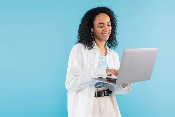 Brunetta donna afroamericana in camicia bianca sorridente durante l'utilizzo di laptop isolato su blu — Foto stock