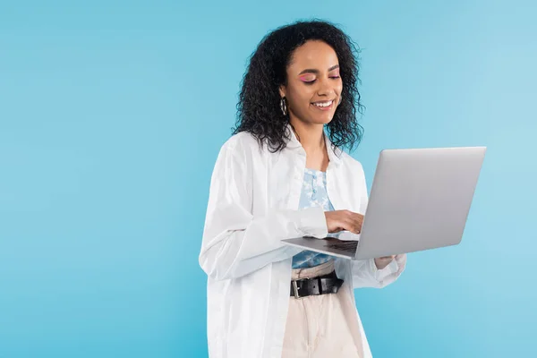 Brunette african american woman in white shirt smiling while using laptop isolated on blue — Stock Photo