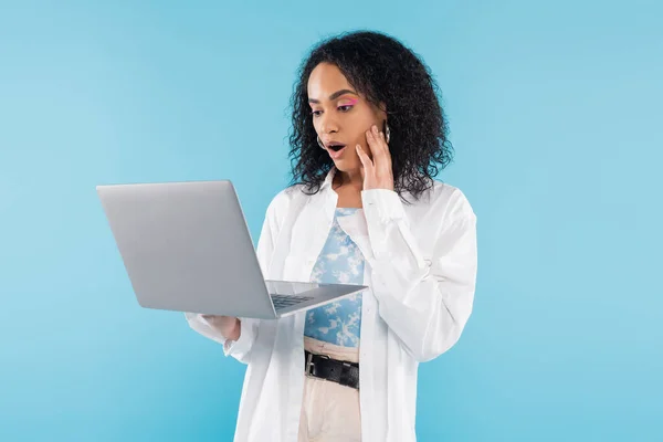 Mujer afroamericana asombrada en camisa blanca mirando el ordenador portátil aislado en azul - foto de stock