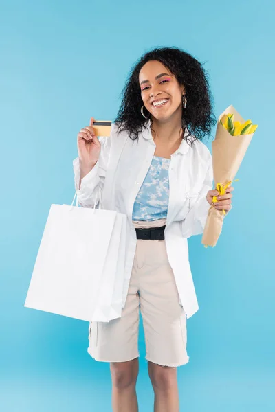 Freudige afrikanisch-amerikanische Frau mit Einkaufstaschen und gelben Tulpen, die Kreditkarte isoliert auf blauem Grund zeigt — Stockfoto