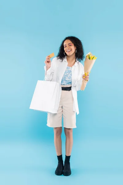 Full length of cheerful african american woman with yellow tulips and shopping bags showing credit card on blue background — Stock Photo