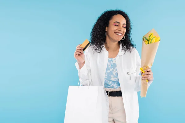 Piacere donna afroamericana con carta di credito e borse della spesa guardando tulipani gialli isolati su blu — Foto stock