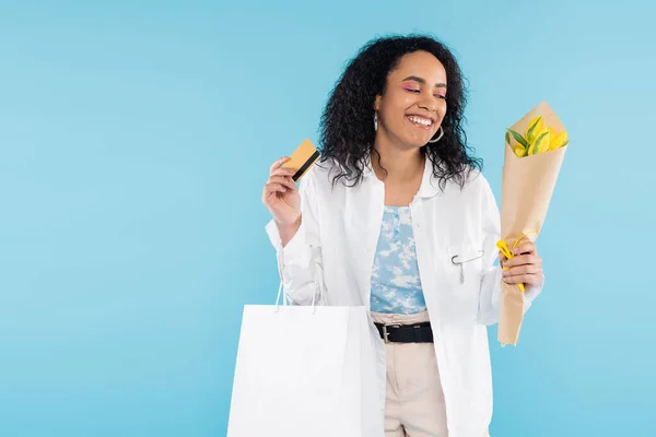 Pleased african american woman with credit card and shopping bags looking at yellow tulips isolated on blue — Stock Photo