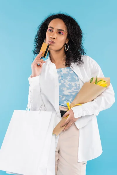 Pensive african american woman with purchases and yellow tulips looking away isolated on blue — Stock Photo