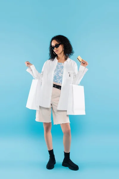 Longitud completa de la elegante mujer afroamericana en pantalones cortos blancos y gafas de sol posando con tarjeta de crédito y bolsas de compras sobre fondo azul - foto de stock