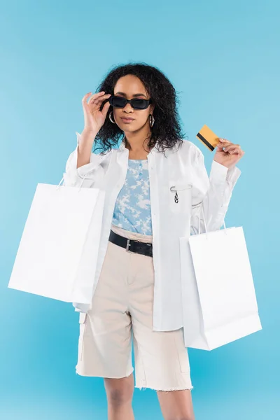 Femme afro-américaine avec sacs à provisions et carte de crédit ajustement lunettes de soleil élégantes isolées sur bleu — Photo de stock
