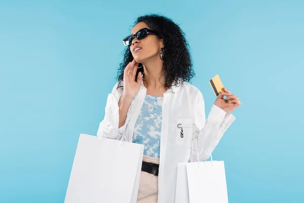 Mujer afroamericana de moda con bolsas de compras y tarjeta de crédito posando en gafas de sol aisladas en azul - foto de stock