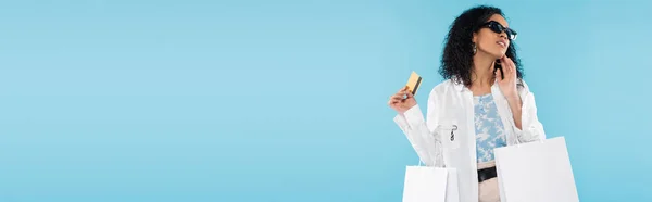 Donna afroamericana in occhiali da sole alla moda in posa con carta di credito e borse della spesa isolate su blu, banner — Foto stock