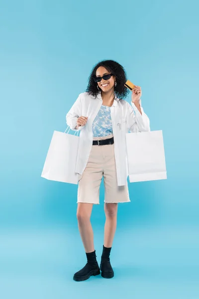 Полная длина модной африканской американки в солнцезащитных очках позирует с белыми пакетами покупок и кредитной картой на синем фоне — стоковое фото