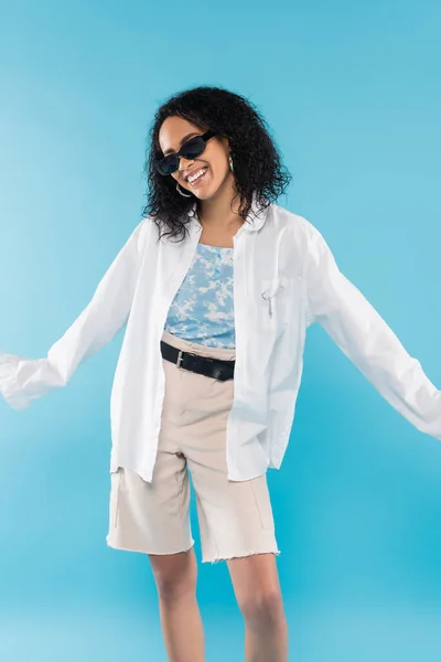 Приємна афро-американська жінка в білій сорочці і сонцезахисних окулярах з розпростертими руками ізольованими на синьому — стокове фото