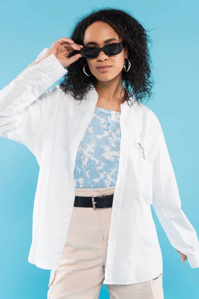 Fashionable african american woman in white shirt adjusting sunglasses isolated on blue — Stock Photo