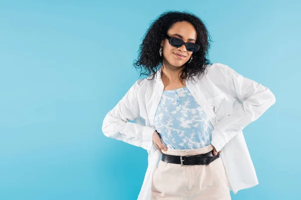 Bruna donna afroamericana in camicia bianca ed eleganti occhiali da sole in piedi con le mani sulla vita isolate sul blu — Foto stock