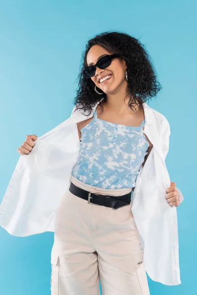 Allegra donna afro americana in posa in occhiali da sole e camicia bianca elegante isolato su blu — Foto stock