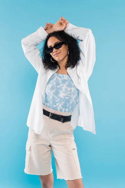 Brunette african american woman in trendy sunglasses and white shirt standing with hands above head isolated on blue — Stock Photo