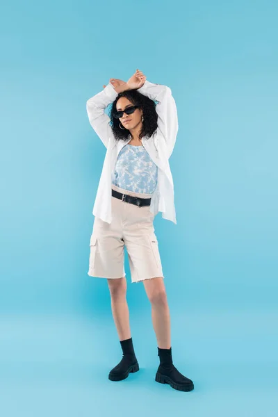 Full length of young african american woman in shorts and sunglasses posing with hands above head on blue background — Stock Photo