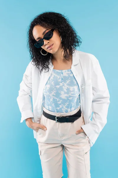 Брюнетка африканская американка в модных солнцезащитных очках позирует с руками в карманах хлопковых шорт, изолированных на голубом — стоковое фото