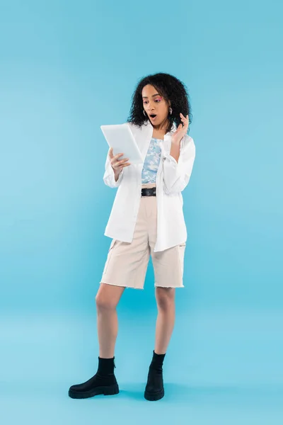 Pleine longueur de femme afro-américaine étonnée dans des vêtements à la mode en regardant tablette numérique sur fond bleu — Photo de stock